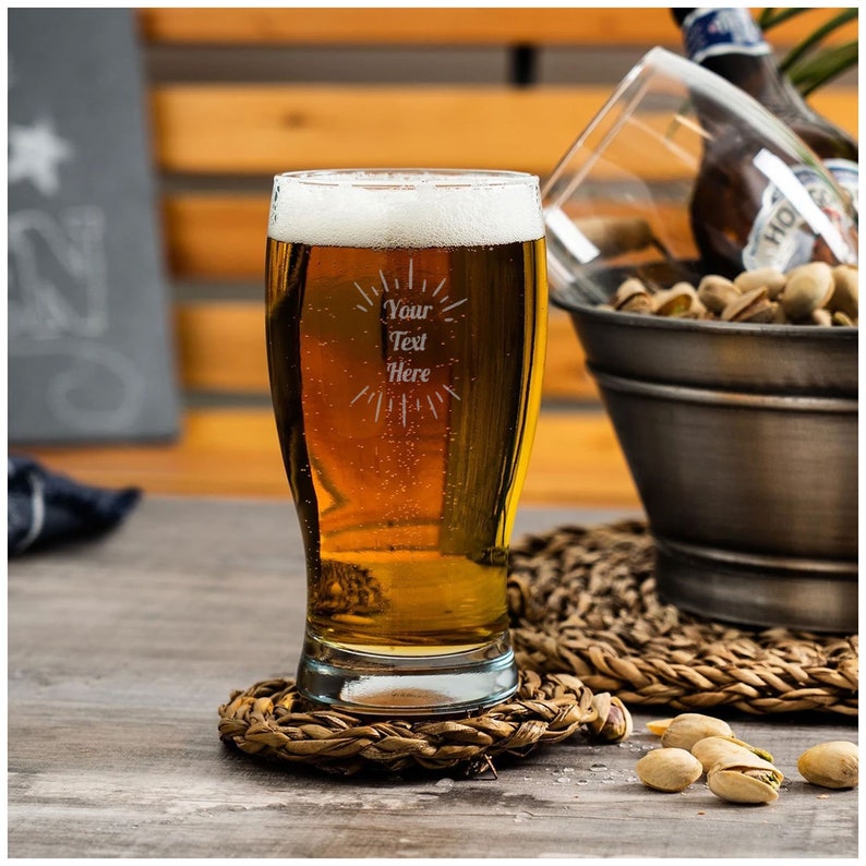 May include: A clear glass beer mug with a frosted design that says "Your Text Here". The mug is filled with a golden beer and is sitting on a woven coaster.