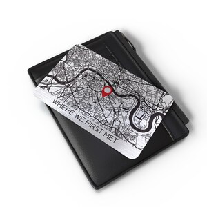 Puede incluir: Una tarjeta de metal plateado con un mapa en blanco y negro de una ciudad. Un pin rojo est&aacute; colocado en el mapa. La tarjeta tiene el texto "WHERE WE FIRST MET" impreso en ella. La tarjeta est&aacute; encima de una cartera de cuero negro.