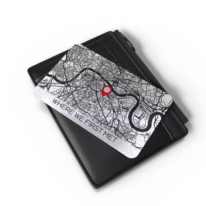 Puede incluir: Una tarjeta de metal plateado con un mapa en blanco y negro de una ciudad. Un pin rojo est&aacute; colocado en el mapa. La tarjeta tiene el texto "WHERE WE FIRST MET" impreso en ella. La tarjeta est&aacute; encima de una cartera de cuero negro.