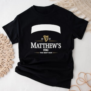 May include: Black t-shirt with a white banner and gold harp graphic. The text reads "Matthew's Stag" with "The Best Man" below. The shirt also has the numbers "20" and "25" on the design.