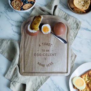 May include: A wooden serving tray shaped like a slice of toast with two egg cups and the text "A Toast to an Egg-Celent Name Here".