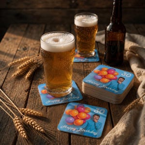 Può includere: Due bicchieri di birra dorata con schiuma, una bottiglia di birra in vetro scuro e una pila di sottobicchieri quadrati. Ogni sottobicchiere presenta un grafico di una persona che tiene un mazzo di palloncini colorati con il testo Happy Birthday e il numero 30.