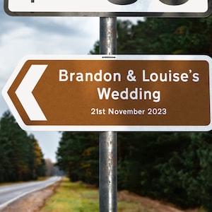 May include: A brown and white directional sign with a white arrow pointing left. The sign reads "Brandon & Louise's Wedding 21st November 2023".
