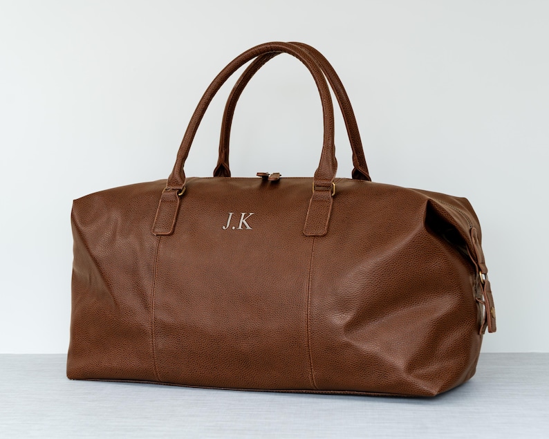 May include: A brown leather duffel bag with a zipper closure and two top handles. The bag has the initials "J.K." embossed on the front.