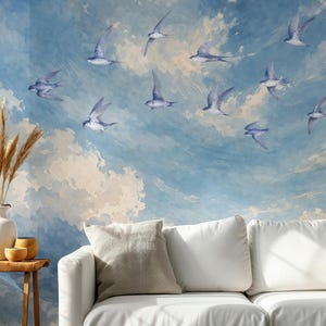 May include: A wall mural featuring a watercolor-style blue sky with white clouds and a flock of birds in flight. The scene is positioned behind a white sofa, a wooden side table, a white vase with dried grasses, and a small wooden bowl.