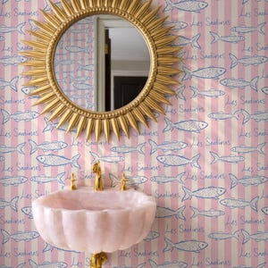 May include: A bathroom scene featuring a gold sunburst mirror above a pink, scalloped sink with gold fixtures. The wallpaper has a pink and white striped pattern with blue fish illustrations and the text "Les Sardines".