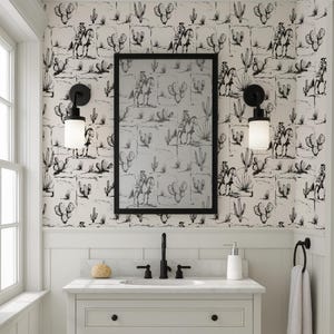 May include: Bathroom with a black and white cowboy and cactus wallpaper. A black framed mirror hangs above a white vanity with a black faucet and sconces. A white towel hangs on a black towel ring.
