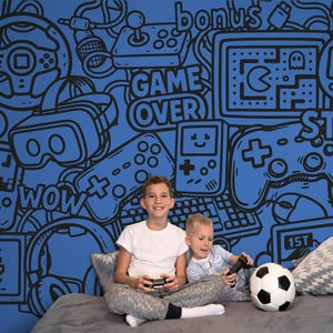 Puede incluir: Dos niños sentados en una cama con un balón de fútbol, sosteniendo mandos de videojuegos. La pared azul detrás de ellos presenta un diseño de garabatos en blanco y negro de elementos de videojuegos, incluyendo mandos y la frase "GAME OVER".