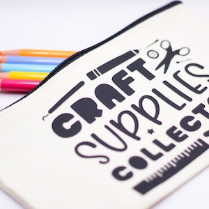 May include: A cream-coloured pencil case with the text "Craft Supplies Collector" in black. The case is open, revealing several coloured pencils in pink, yellow, blue, and orange. A black zip runs along the top.