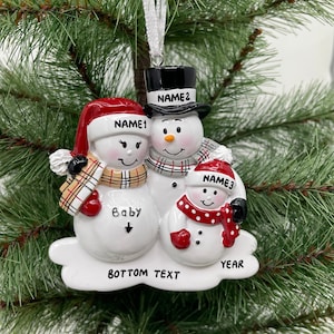 May include: White snowman family Christmas ornament with red, black and white hats. The snowman on the left is pregnant and has a plaid scarf. The snowman on the right is holding a red and white polka dot scarf. The ornament says "Baby" with an arrow pointing down, "Bottom Text", and "Year".
