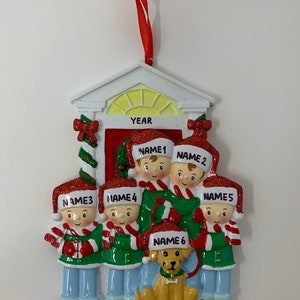 May include: A white and red Christmas ornament depicting a family of six standing in front of a house with a dog. The ornament has the words "YEAR", "FAMILY NAME", and the names of each family member.