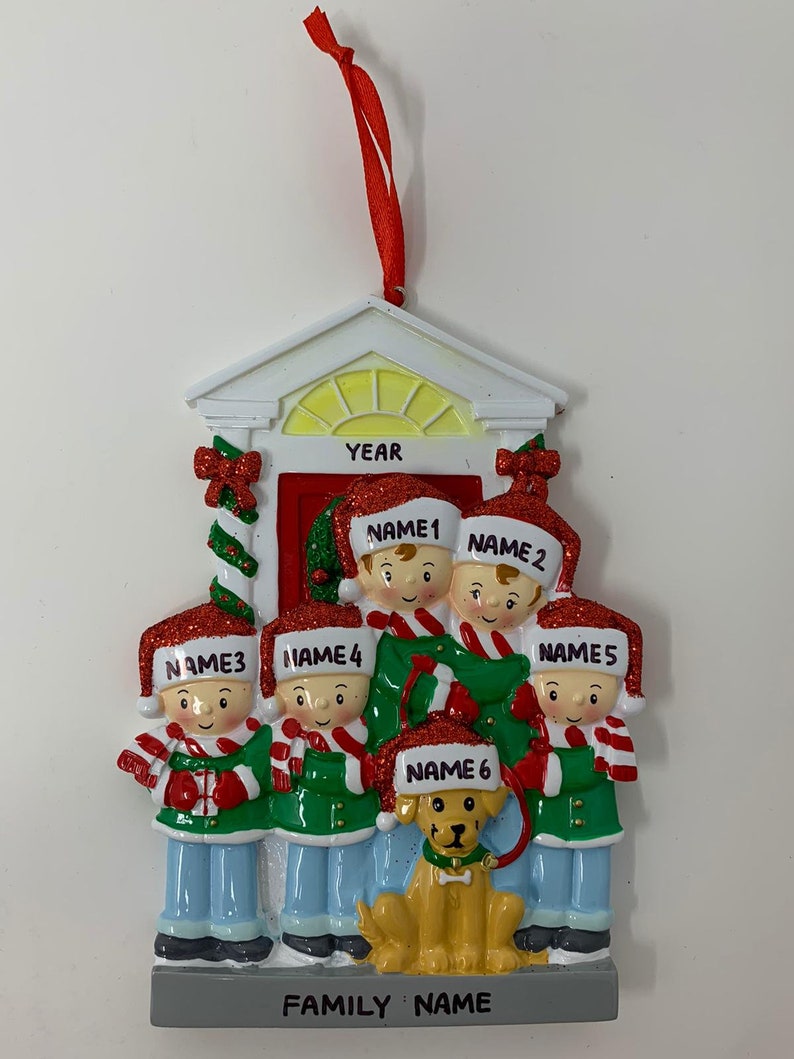 May include: A white and red Christmas ornament depicting a family of six standing in front of a house with a dog. The ornament has the words "YEAR", "FAMILY NAME", and the names of each family member.