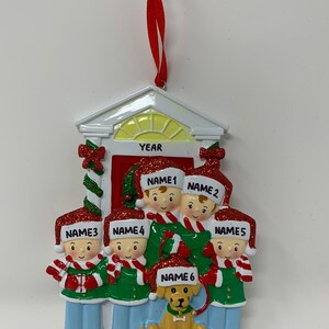 May include: A white Christmas ornament shaped like a house with a red door and six figures standing in front of it. The figures are wearing red and green hats and scarves. The text "FAMILY NAME" is printed on the bottom of the ornament. The text "YEAR" is printed above the door.