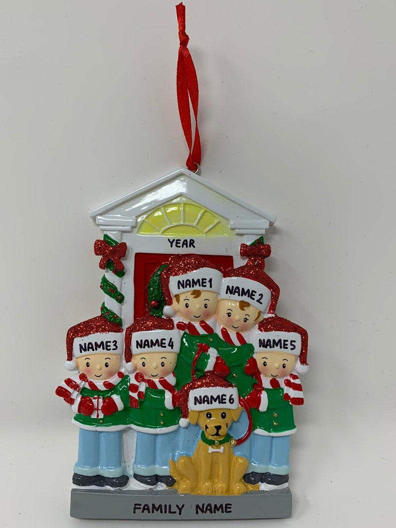 May include: A white Christmas ornament shaped like a house with a red door and six figures standing in front of it. The figures are wearing red and green hats and scarves. The text "FAMILY NAME" is printed on the bottom of the ornament. The text "YEAR" is printed above the door.