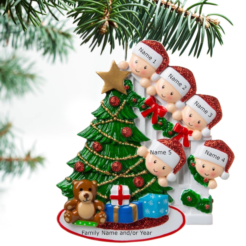 May include: A white ceramic Christmas ornament depicting a family of five peeking over a white fence, with a green Christmas tree, a brown teddy bear, and wrapped presents. The ornament is decorated with red and green glitter. The text "Family Name and/or Year" is printed on the bottom.