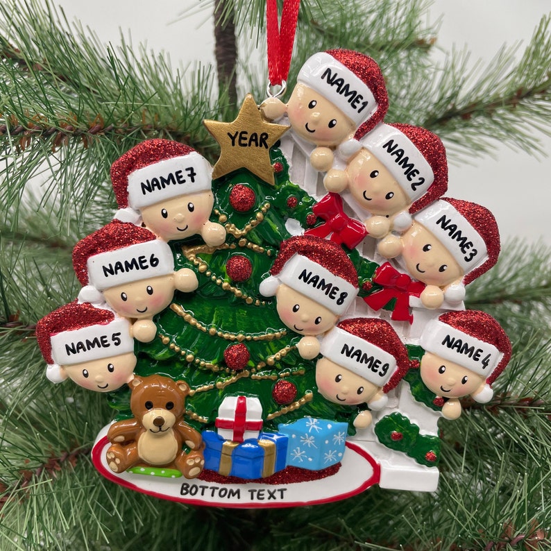 May include: A white ceramic Christmas tree ornament with nine red-hatted figures peeking around the tree. The ornament features a green Christmas tree with a gold star, a teddy bear, and a blue gift box. The text "YEAR" is on the star and "BOTTOM TEXT" is at the bottom of the ornament.