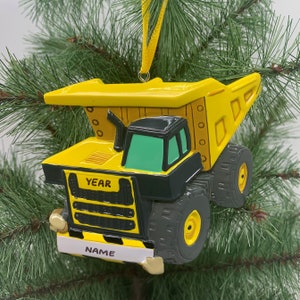 Puede incluir: Adorno de Navidad de cami&oacute;n volquete de juguete amarillo con detalles negros. El cami&oacute;n tiene una cabina negra con un parabrisas verde y las palabras "YEAR" y "NAME" en la parte delantera.