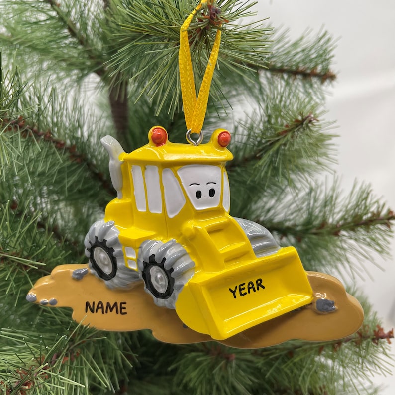 Puede incluir: Adorno de Navidad de veh&iacute;culo de construcci&oacute;n de juguete amarillo con una cara sonriente. El adorno est&aacute; colgado de una cinta amarilla y tiene las palabras "NAME" y "YEAR" en &eacute;l.