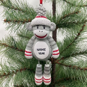 May include: A gray sock monkey Christmas ornament with a white pom-pom hat, red and white striped cuffs, and a white circle with the words "NAME YEAR" on the front.