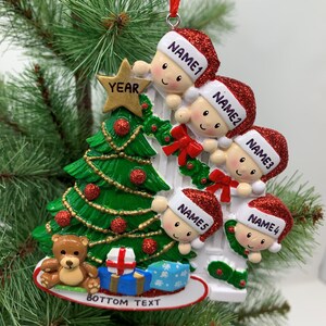 May include: A white ceramic Christmas ornament featuring five cartoon children peeking over a white picket fence. The children are wearing red Santa hats with glitter and have smiling faces. A green Christmas tree with red and gold ornaments is behind the fence. The ornament also includes a teddy bear, a wrapped gift, and the text "YEAR" and "BOTTOM TEXT".