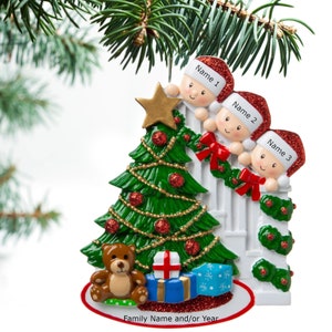 May include: A Christmas ornament featuring three children peeking over a white picket fence, decorated with red bows and green garland. A green Christmas tree with red ornaments and a gold star sits in front of the fence. A brown teddy bear and wrapped presents are at the base of the tree. The ornament reads "Family Name and/or Year".