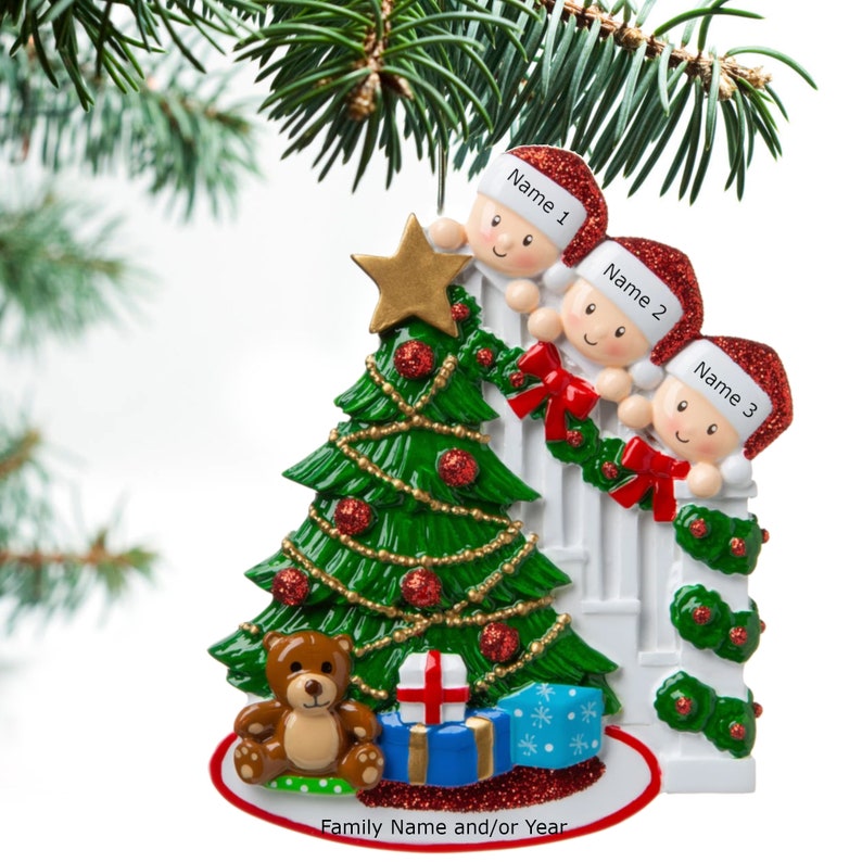 May include: A Christmas ornament featuring three children peeking over a white picket fence, decorated with red bows and green garland. A green Christmas tree with red ornaments and a gold star sits in front of the fence. A brown teddy bear and wrapped presents are at the base of the tree. The ornament reads "Family Name and/or Year".