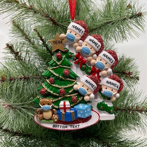 May include: A white Christmas tree ornament with four figures wearing Santa hats and face masks. The figures are peeking over a white picket fence. The ornament also features a small teddy bear wearing a face mask, a star, and presents. The text "YEAR" is on the star. The text "BOTTOM TEXT" is on the base of the ornament.