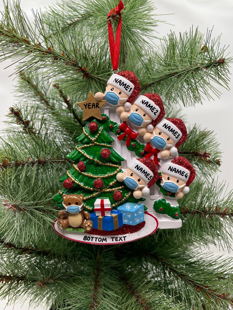 May include: A white Christmas tree ornament with four figures wearing Santa hats and face masks. The figures are peeking over a white picket fence. The ornament also features a small teddy bear wearing a face mask, a star, and presents. The text "YEAR" is on the star. The text "BOTTOM TEXT" is on the base of the ornament.
