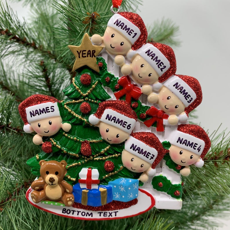 May include: A white Christmas tree ornament with six children peeking out from behind it. The children are wearing red Santa hats with white trim and have smiling faces. The tree is decorated with a gold star, red and green ornaments, and a red bow. The ornament also features a teddy bear, a blue gift box, and the text "BOTTOM TEXT".