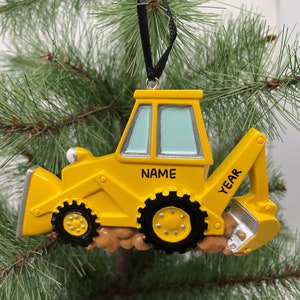 Puede incluir: Adorno de Navidad de excavadora amarilla de juguete con una cinta negra. El adorno tiene espacio para un nombre y un a&ntilde;o.