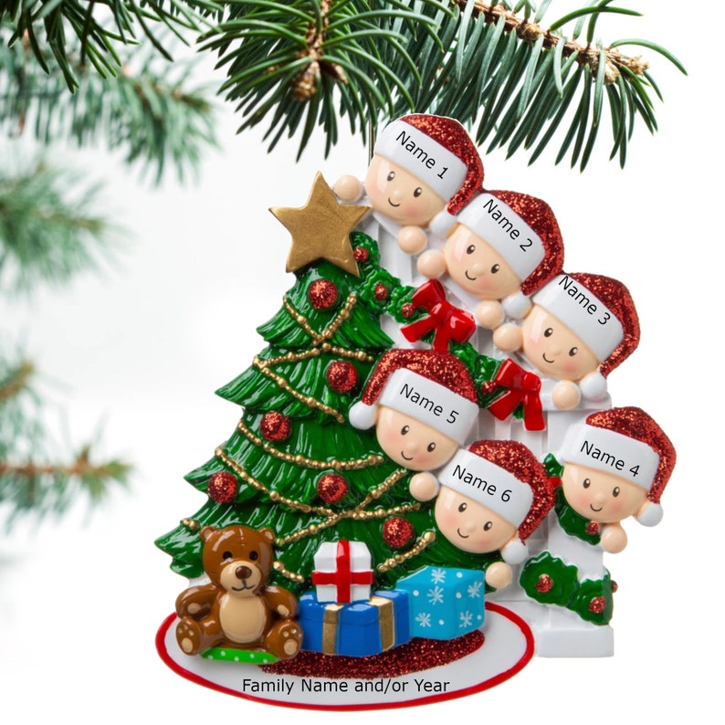 May include: A white ceramic Christmas tree ornament with six figures peeking over a fence. The figures are wearing red Santa hats and have personalized names. The tree is decorated with red and gold ornaments. A teddy bear and presents are at the base of the tree. The ornament reads "Family Name and/or Year".