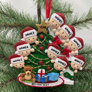 May include: A white Christmas tree ornament with nine red-hatted figures peeking out from behind the tree. The figures have names written on their hats, and the tree has a gold star on top with the word "YEAR" written on it. The ornament also features a teddy bear, a wrapped gift, and the text "BOTTOM TEXT" at the bottom.