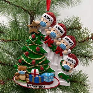 May include: A Christmas ornament featuring a family of four wearing Santa hats and face masks, decorating a Christmas tree. The ornament also includes a teddy bear wearing a face mask and a banner that says "BOTTOM TEXT".