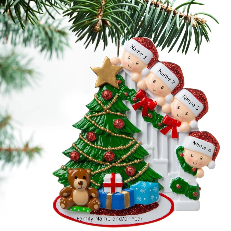 May include: A white ceramic Christmas ornament featuring four children peeking over a white picket fence, a green Christmas tree with red ornaments, a teddy bear, and a wrapped gift. The ornament has a space for a family name and/or year.