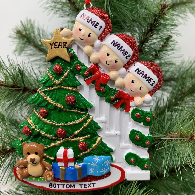 May include: A white Christmas tree ornament with three children peeking over a white picket fence. The children are wearing red Santa hats with their names, "NAME1", "NAME2", and "NAME3", written on them. The ornament also features a green Christmas tree with red ornaments, a gold star, a brown teddy bear, and wrapped presents. The bottom of the ornament says "BOTTOM TEXT" and "YEAR".