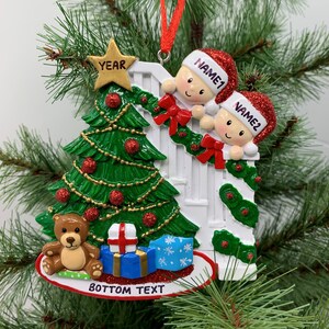 May include: A white Christmas ornament with a green Christmas tree, two children peeking over a white picket fence, a teddy bear, and a blue gift box. The ornament has the text "YEAR" on a gold star, "NAME1" and "NAME2" on the children's hats, and "BOTTOM TEXT" at the bottom.