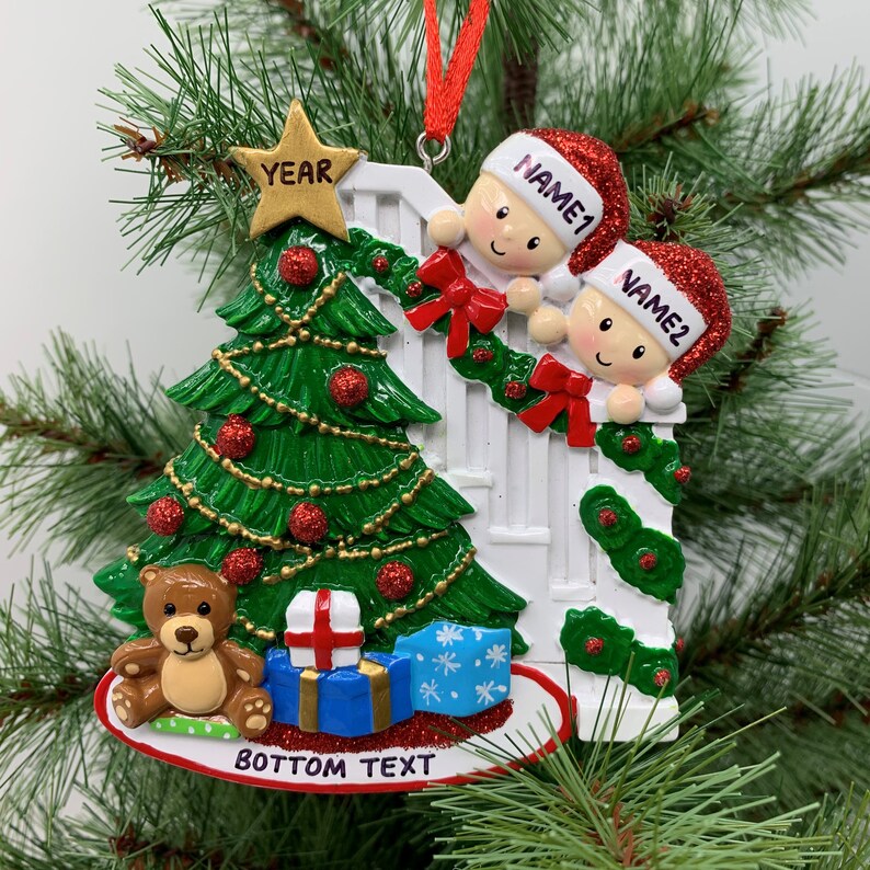 May include: A white Christmas ornament with a green Christmas tree, two children peeking over a white picket fence, a teddy bear, and a blue gift box. The ornament has the text "YEAR" on a gold star, "NAME1" and "NAME2" on the children's hats, and "BOTTOM TEXT" at the bottom.