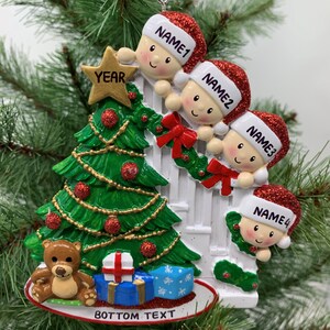May include: A white Christmas ornament with four children peeking over a white picket fence. The children are wearing red Santa hats and have smiling faces. A green Christmas tree with red and gold ornaments is in the background. The ornament also features a teddy bear, a wrapped present, and the text "YEAR" and "BOTTOM TEXT".