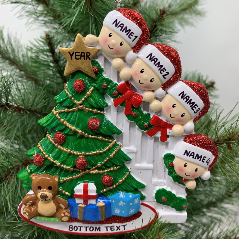 May include: A white Christmas ornament with four children peeking over a white picket fence. The children are wearing red Santa hats and have smiling faces. A green Christmas tree with red and gold ornaments is in the background. The ornament also features a teddy bear, a wrapped present, and the text "YEAR" and "BOTTOM TEXT".