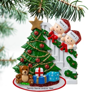 May include: A white picket fence Christmas ornament with two children peeking over the top. The children are wearing red Santa hats and have smiling faces. A green Christmas tree with red ornaments and a gold star on top is in the background. There is a brown teddy bear and wrapped presents at the base of the tree. The ornament has a white circle at the bottom with the text "Family Name and/or Year".