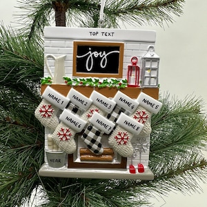 May include: White ceramic Christmas ornament depicting a fireplace with stockings hanging from the mantel. The stockings are personalized with names and the fireplace has a chalkboard with the word "joy" written on it.