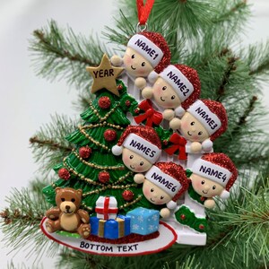 May include: A white Christmas tree ornament with six children peeking out from behind a white picket fence. The children are wearing red Santa hats and have names written on them. The ornament also features a green Christmas tree with red ornaments, a teddy bear, and presents. The bottom of the ornament says "BOTTOM TEXT" and the top says "YEAR".