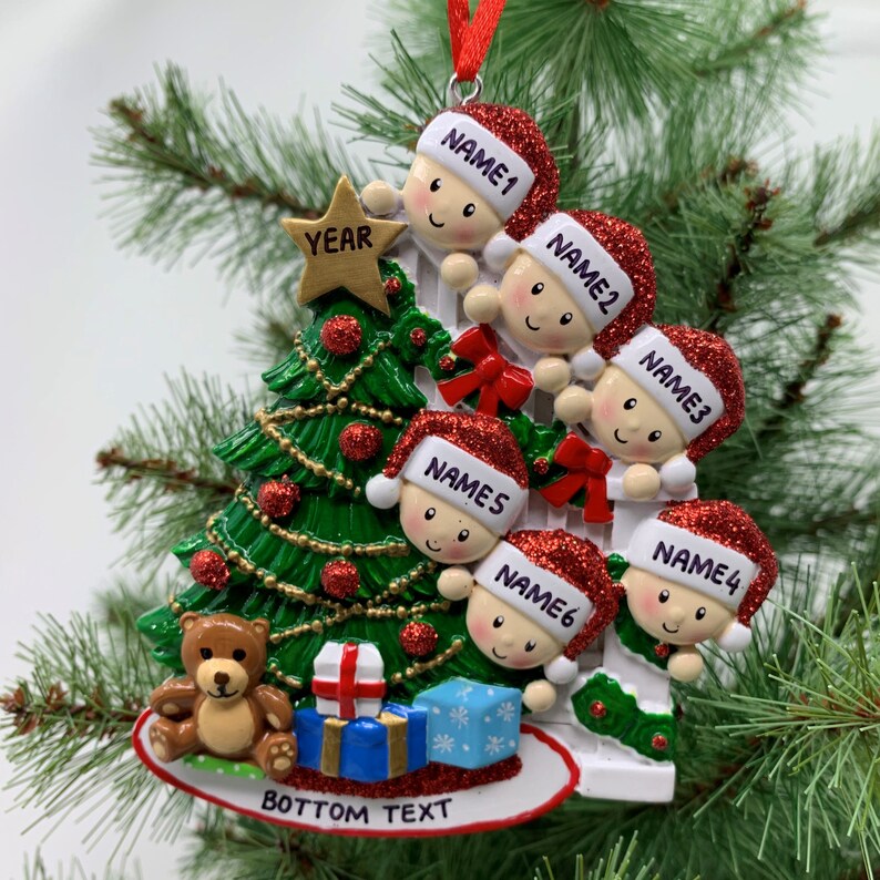 May include: A white Christmas tree ornament with six children peeking out from behind a white picket fence. The children are wearing red Santa hats and have names written on them. The ornament also features a green Christmas tree with red ornaments, a teddy bear, and presents. The bottom of the ornament says "BOTTOM TEXT" and the top says "YEAR".
