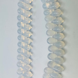 Könnte beinhalten: Eine Halskette aus weißen Opalperlen mit einem silbernen Verschluss. Die Perlen sind tropfenförmig und auf einem durchsichtigen Faden aufgefädelt.