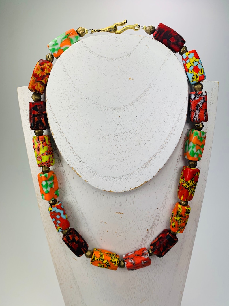 May include: A colorful necklace featuring rectangular beads in shades of red, orange, yellow, and green. The beads are strung together with small brass-colored spacers and a brass clasp. The necklace is displayed on a white bust.