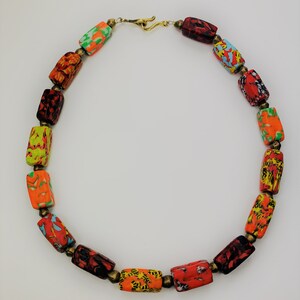 May include: A colorful necklace featuring rectangular beads with vibrant patterns in red, orange, yellow, green, and blue. The beads are strung together with small brass-colored connectors and a gold-tone clasp.