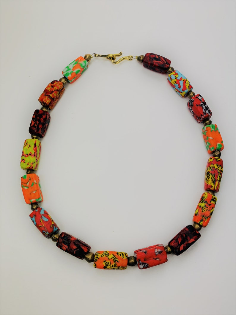 May include: A colorful necklace featuring rectangular beads with vibrant patterns in red, orange, yellow, green, and blue. The beads are strung together with small brass-colored connectors and a gold-tone clasp.