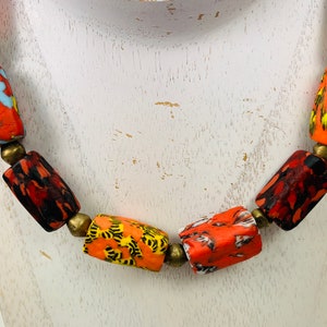 May include: A colorful necklace featuring cylindrical beads in shades of red, orange, yellow, and green. The beads have a mottled pattern and are separated by small, brass-colored spacers. The necklace is displayed on a white surface.
