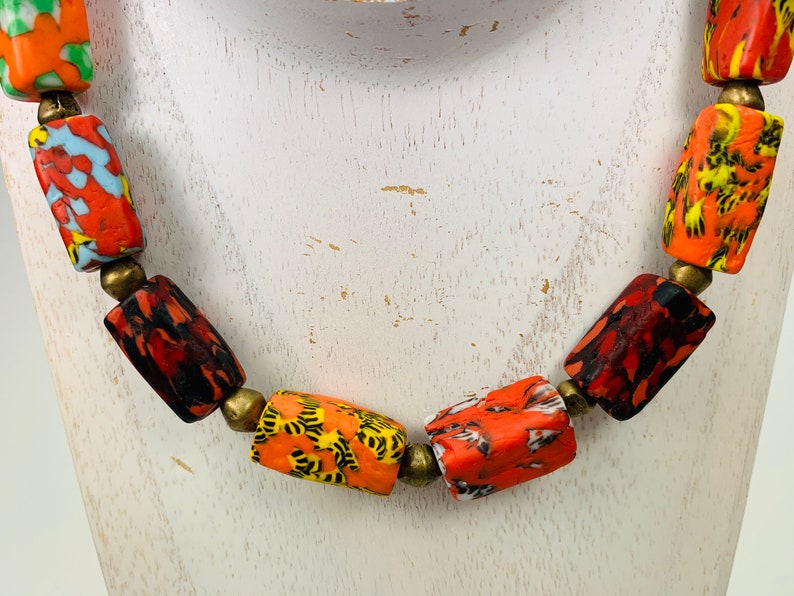 May include: A colorful necklace featuring cylindrical beads in shades of red, orange, yellow, and green. The beads have a mottled pattern and are separated by small, brass-colored spacers. The necklace is displayed on a white surface.