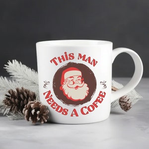 May include: White ceramic mug with a red and brown illustration of Santa Claus and the text "This Man Needs A Coffee".